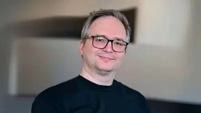 Alexander trägt ein schwarzes T-Shirt und eine Brille und lächelt in die Kamera. 