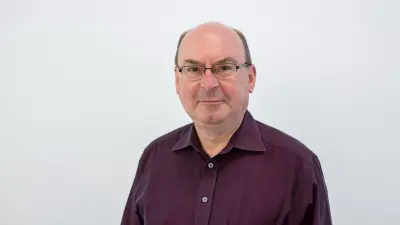 Franz trägt ein brombeerfarbenes Hemd und eine Brille und lächelt in die Kamera.