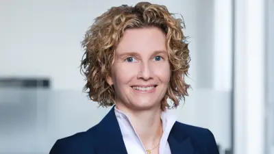 Nicolet trägt einen Blazer, eine Bluse und lockige Haare – sie lächelt in die Kamera.