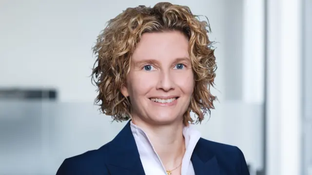 Nicolet trägt einen Blazer, eine Bluse und lockige Haare – sie lächelt in die Kamera.