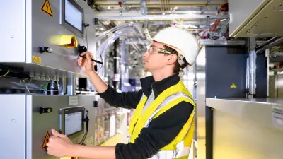 Person mit Schutzhelm und Warnweste arbeitet an Steuerungseinheit in technischer Umgebung