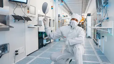 Ein Mitarbeiter der Halbleiterfertigung im Reinraumanzug balanciert einen Wafer auf seinem Zeigefinger.
