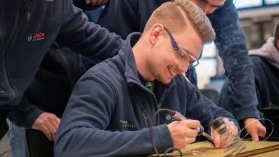 Ein Auszubildender lötet konzentriert eine Platine, während zwei Kollegen ihm aufmerksam über die Schulter schauen. 