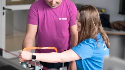 Eine Auszubildende arbeitet mit einer Handsäge an einem Werkstück, während ein Ausbilder sie dabei freundlich unterstützt. Beide lächeln, die Stimmung ist motivierend und kollegial.