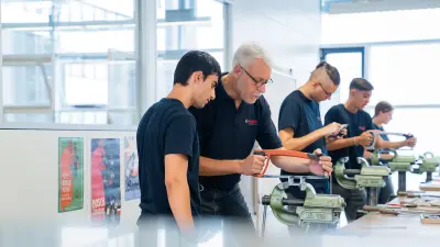 Ein Mechanik-Ausbilder steht mit drei Auszubildenden an einer Werkbank und unterstützt sie beim korrekten Sägen von Metall.