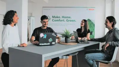 Vier junge Menschen lachen zusammen am Tisch im Bosch-Büro mit Laptops und grünem Poster im Hintergrund.