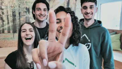 Vier junge Menschen lachen in die Kamera, eine zeigt ein Peace-Zeichen, Hintergrund mit Waldmotiv.