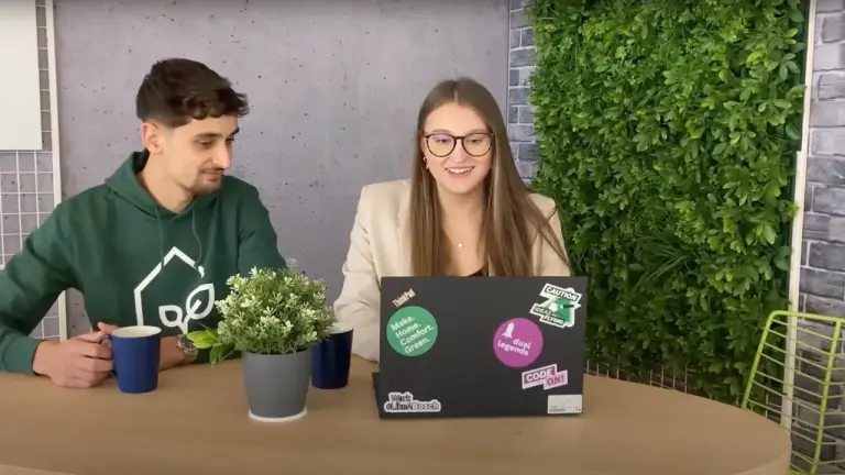 Zwei junge Menschen arbeiten gemeinsam an einem Laptop mit Stickern, grüne Pflanzen im Hintergrund.