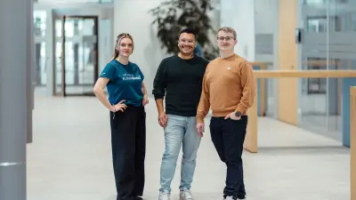 Drei Personen stehen in einem modernen Bürogebäude, schauen in die Kamera und lächeln.