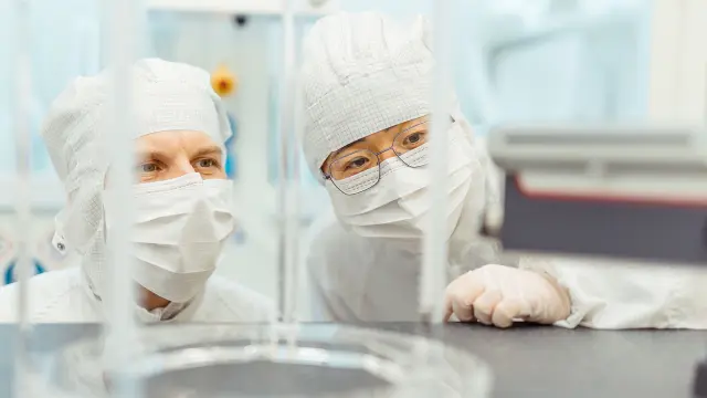Zwei Personen in weißer Schutzkleidung blicken über einen Labortisch mit Glasgefäßen.