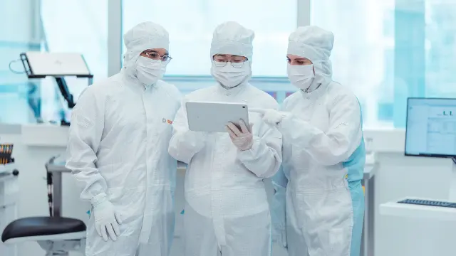 Drei Personen in weißen Schutzanzügen stehen in einem Labor und betrachten gemeinsam ein Tablet.
