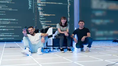 Drei Auszubildende sitzen vor einer IT-Wand auf dem Boden und schauen in die Kamera