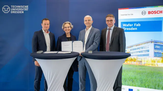 Three men and a woman proudly present a document, with a banner of the Bosch Wafer Fab Dresden and the logo of the Technical University of Dresden in the background.