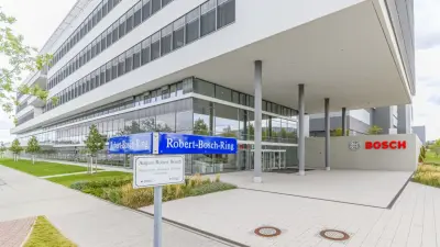 Der Eingang zum vierstöckigen, modernen Gebäude am Bosch-Standort in Dresden mit einem großen, überdachten Außenbereich. Davor sieht man das Straßenschild, das in beide Richtungen den Robert-Bosch-Ring anzeigt.