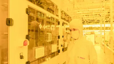 Ein Mensch in weißer Schutzkleidung mit Bosch Logo schaut in einer Industriehalle in einen Schaltschrank. Das Gelblicht dient dazu, die empfindlichen Prozesse und Materialien vor schädlicher UV-Strahlung zu schützen.