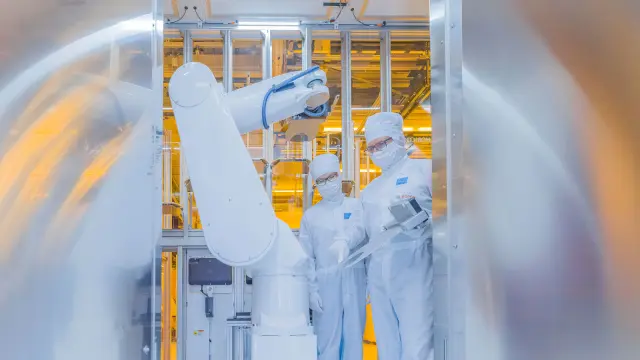 Hinter einer offenen Glastüre inspizieren zwei Mitarbeitende in Schutzkleidung einen Roboterarm.