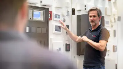 Ein Mitarbeiter erklärt etwas vor einer Wand mit Panels, Alarmsystemen und anderen Bosch Building Technologies Produkten.