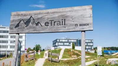 Ein hölzernes Schild auf dem Berge und die Beschriftung "eTrail" zu sehen sind befindet sich am Eingang zu einer Teststrecke für eBikes. Im Hintergrund sind einige Bürogebäude zu sehen.