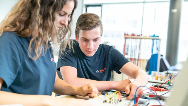 Zwei Auszubildene arbeiten gemeinsam an einer Elektronikschaltung in einem technischen Arbeitsumfeld.