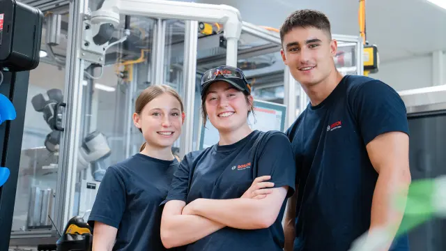 Ein Team bestehend aus drei Auszubildenden des Bosch-Standorts Feuerbach steht vor einem Roboter. Alle drei lachen und tragen Arbeitskleidung, die Auszubildende in der Mitte trägt eine Kappe und Schutzbrille auf dem Kopf und verschränkt die Arme.