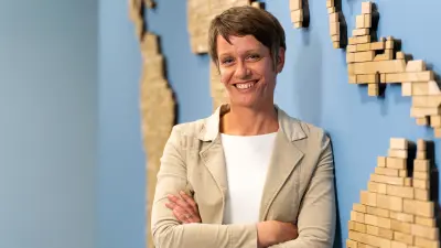 Corinna Dornbusch steht angelehnt an einer blauen Wand mit Holzelementen, trägt kurze Haare, ein weißes T-Shirt und einen beigen Blazer und lächelt in die Kamera.