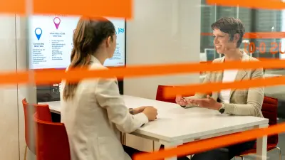 In einem Konferenzraum sitzt Corinna Dornbusch einer Kollegin gegenüber, mit der sie spricht. Man sieht orangene Klebestreifen auf der Fensterfront des Raumes.