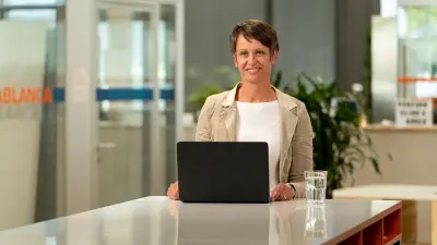 Corinna Dornbusch sitzt an einem Tisch mit einem Laptop und einem Glas Wasser vor ihr und lächelt in die Kamera.