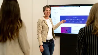 Corinna Dornbusch steht vor einem Bildschirm, auf dem Infos zu agilen HR Lösungen zu sehen sind und deutet darauf. Sie sieht zwei Kolleginnen an, die vor ihr stehen.