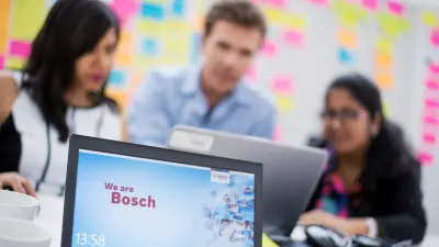 Drei Personen sitzen im Hintergrund. Sie sind unscharf. Im Vordergrund steht ein aufgeklappter Laptop auf einem Tisch.