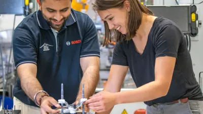 Chiara prüft gemeinsam mit einem Kollegen ein Maschinenbauteil auf Funktionstüchtigkeit in der Fertigungshalle. Sie lachen.