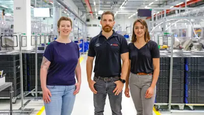 Verena (links), Kevin (Mitte) und Chiara (rechts) stehen in einem Gang der Fertigungshalle. Im Hintergrund sind Maschinen und Rollcontainer zu sehen.