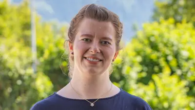 Verena steht lächelnd vor Bäumen und blickt in die Kamera. Sie ist bis zum Oberkörper zu sehen.