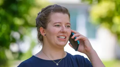 Verena steht draußen zwischen Bäumen und telefoniert lachend.