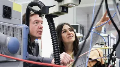 Rolf und Hande befinden sich in einer Werkstatt-Labor-Umgebung vor technischen Arbeitsgeräten, die verkabelt sind. Hande deutet mit ihrer linken Hand auf etwas, Rolf hat ebenfalls einen Arm nach vorne gestreckt und folgt ihr mit seinem Blick.