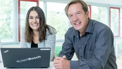 Hande ist links im Bild und lehnt mit ihrem Oberkörper auf einem Tisch und lächelt in die Kamera. Vor ihr steht ein Laptop. Rechts neben ihr ist Rolf zu sehen, der ebenfalls mit dem Oberkörber auf dem Tisch lehnt und in die Kamera lächelt. Er hat seine Hände vor sich gefaltet.