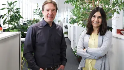 Rolf und Hande sind in einer Büroumgebung zu sehen. Rolf steht links im Bild, Hande rechts. Beide lächeln in die Kamera. Im Hintergrund befinden sich grüne Pflanzen, Sideboards und Schreibtische.