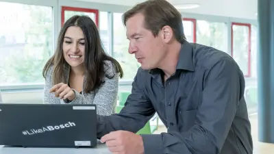 Hande ist links im Bild und lehnt mit ihrem Oberkörper auf einem Tisch und lächelt in Richtung des Laptops, der vor ihr steht. Ihre Hand deutet auf den Bildschirm. Auf dem Laptop ist ein Sticker mit dem Schriftzug #LikeABosch. Rechts neben ihr ist Rolf zu sehen, der ebenfalls mit dem Oberkörber auf dem Tisch lehnt und in Richtung Laptop schaut.