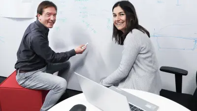Rolf und Hande sitzen vor einem großen Whiteboard und lächeln in die Kamera. Rolf sitzt links und hat einen Stift in der Hand, den er in Richtung Whiteboard hält. Darauf sind viele Notizen und Formeln zu sehen. Im Vordergrund befindet sich ein Tisch, auf dem ein Laptop steht.