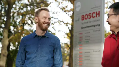 Christoph Dörr steht gemeinsam mit einem Kollegen draußen vor einem Schild, auf dem das Bosch-Logo zu erkennen ist. Sie blicken sich gegenseitig an.
