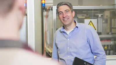Christoph trägt ein blaues Hemd, er blickt in die Kamera und lächelt. Im Hintergrund ist eine Laborumgebung zu sehen.