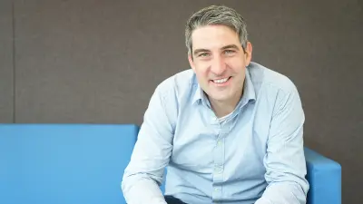 Christoph ist in einer Laborumgebung zu sehen mit einem Laptop in der Hand. Er trägt ein blaues Hemd. Sein Blick ist auf eine Person gerichtet, die angeschnitten im Vordergrund zu sehen ist.