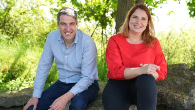Julia und Christoph sitzen in einem Außenbereich nebeneinander auf großen Steinen. Hinter ihnen sind Bäume und Gras zu sehen. Christoph sitzt auf der linken Seite und stützt seine rechte Hand auf seinem rechten Knie ab, er lächelt in die Kamera. Julia hat ihr linkes Bein angewinkelt und stützt ihre Hände darauf ab, sie lächelt in die Kamera.