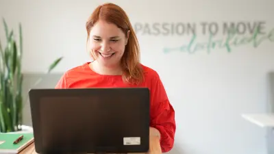 Julia steht an einem Schreibtisch vor einem schwarzen Laptop, ihr Blick ist auf den Laptop gerichtet, sie lächelt. Im Hintergrund ist eine Büroumgebung mit einer Pflanze links zu sehen, rechts ein Schriftzug auf einer Wand, “Passion to move” ist zu lesen.