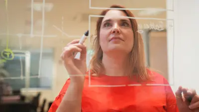 Julia steht vor einer Glasscheibe, auf der ein physikalisches Modell eingezeichnet ist. Sie hält in ihrer rechten Hand einen Stift und setzt zum Zeichnen an. Ihr Blick ist auf die Scheibe gerichtet, ihr Kopf leicht angehoben.