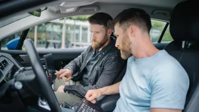 Andreas (vorne) und Dominic (hinten) sitzen auf den Vordersitzen eines Autos. Andreas hat eine Tastatur in der Hand, Dominic zeigt auf einen Bildschirm.