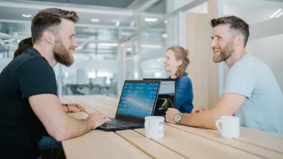 Dominic (links) und Andreas (rechts) sitzen sich an einem Holztisch gegenüber. Beide haben einen Laptop und eine Kaffeetasse vor sich stehen. Sie blicken sich lächelnd an. Im Hintergrund neben Andreas sitzt eine Frau, die ihr Gegenüber ebenfalls anlächelt. 
