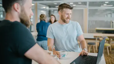 Andreas steht an einem Tisch, vor ihm befinden sich zwei Tassen, ein Glas und ein Laptop. Er blickt lächelnd auf den Bildschirm. Im Vordergrund ist unscharf Dominic zu erkennen, der ebenfalls auf den Bildschirm blickt. Im Hintergrund befinden sich zwei Frauen, die sich auch an einem Tisch gegenüberstehen. Andreas steht an einem Tisch, vor ihm befinden sich zwei Tassen, ein Glas und ein Laptop. Er blickt lächelnd auf den Bildschirm. Im Vordergrund ist unscharf Dominic zu erkennen, der ebenfalls auf den Bildschirm blickt. Im Hintergrund befinden sich zwei Frauen, die sich auch an einem Tisch gegenüberstehen. 