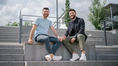 Andreas (links) und Dominic (rechts) sitzen in einem Außenbereich nebeneinander auf einer großen Stufe, die zu einer breiten grauen Treppe gehört. Beide blicken lächelnd in die Kamera.