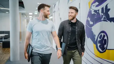 Andreas (links) und Dominic (rechts) laufen einen Gang entlang. Sie blicken sich lächelnd an und haben beide ein Laptop in der Hand. Links von ihnen ist ein Aufenthaltsbereich zu erkennen, rechts eine bunte Wand, auf der ein Fahrzeug illustriert ist.