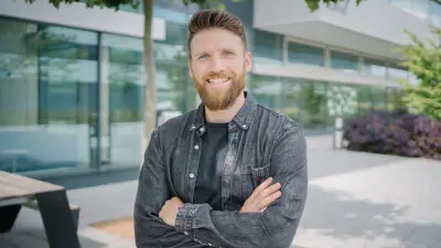 Dominic steht mit verschränkten Armen in einem Außenbereich. Er ist bis zum Oberkörper zu sehen. Er blickt in die Kamera und lächelt.